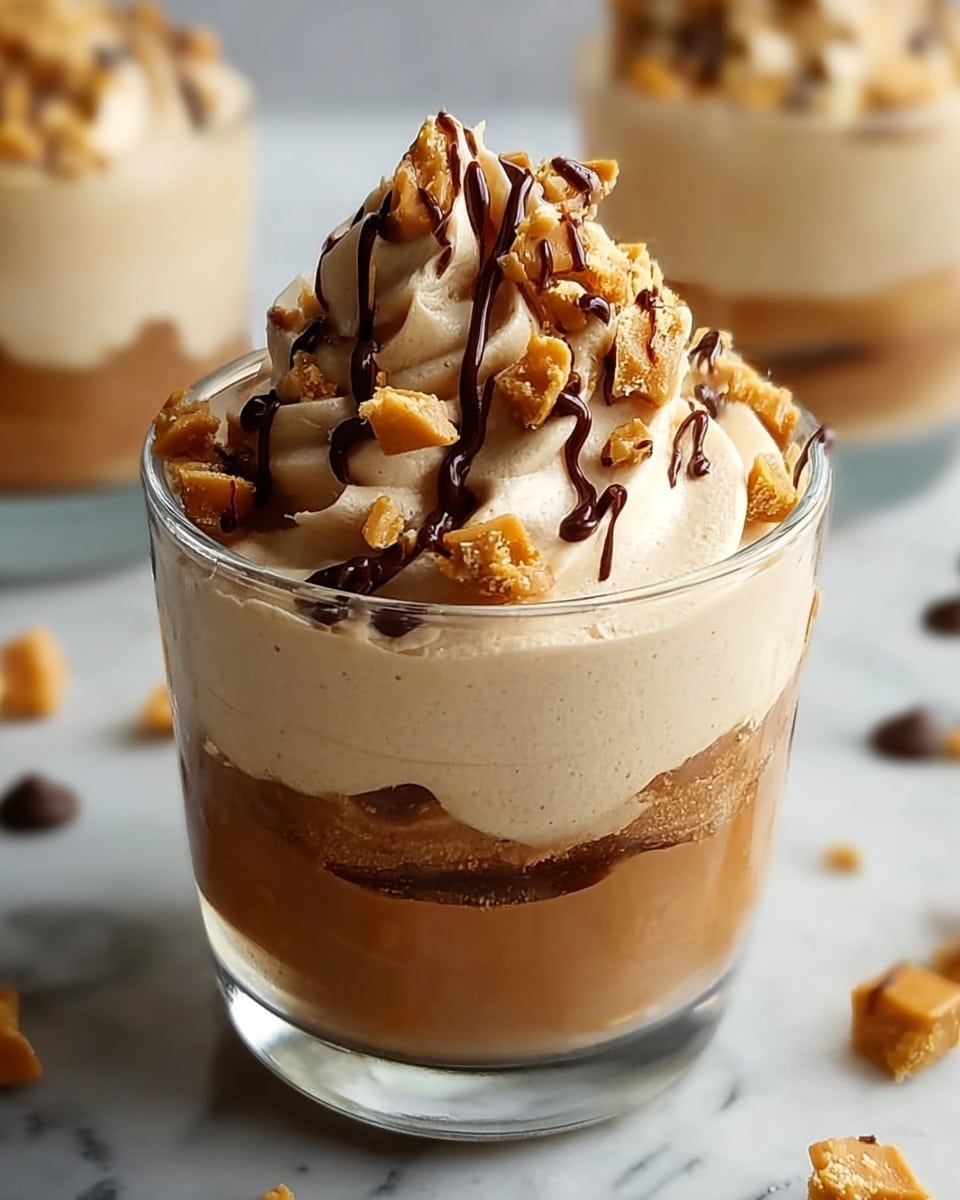 The image shows a clear glass filled with two thick layers of creamy light brown mousse. The bottom layer is smooth and sits just above the base of the glass. The second layer is swirled on top, creating a soft peak that rises above the glass rim. This top layer is garnished with small crunchy pieces that are golden brown, along with a drizzle of dark chocolate sauce running down the sides of the mousse peak. The background is a white marbled surface with blurred elements, including a white bowl filled with more crunchy pieces behind the glass. Photo taken with an iphone --ar 4:5 --v 7