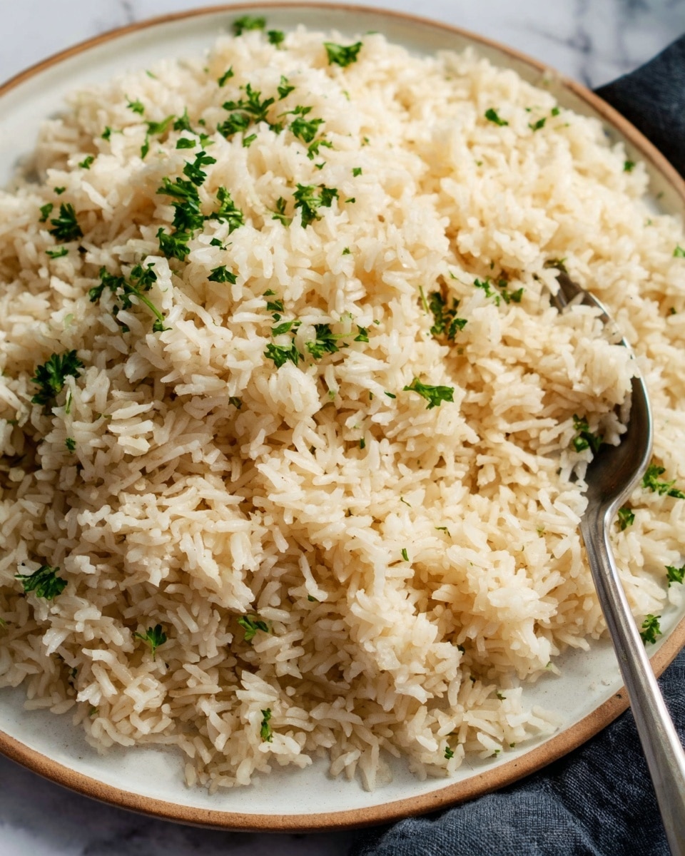 A close-up shot of a large white plate filled with a generous serving of cooked rice, each grain fluffy and slightly separated. The rice is light cream color with small green herb pieces sprinkled evenly on top, adding a touch of freshness and contrast. A gold fork with a rounded handle rests on the edge of the plate, with a woman's hand gently holding it. The background features a white marbled surface, giving a clean and elegant look. photo taken with an iphone --ar 4:5 --v 7