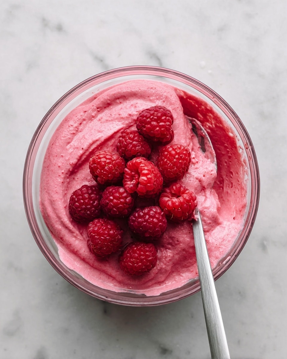 A clear glass bowl filled with a thick, bright pink mixture with a smooth, creamy texture, layered with fresh whole red raspberries sitting on top in the center. A metal spoon coated with the same pink mixture rests inside the bowl, partly submerged in the mixture on the right side. The bowl is placed on a white marbled surface. photo taken with an iphone --ar 4:5 --v 7