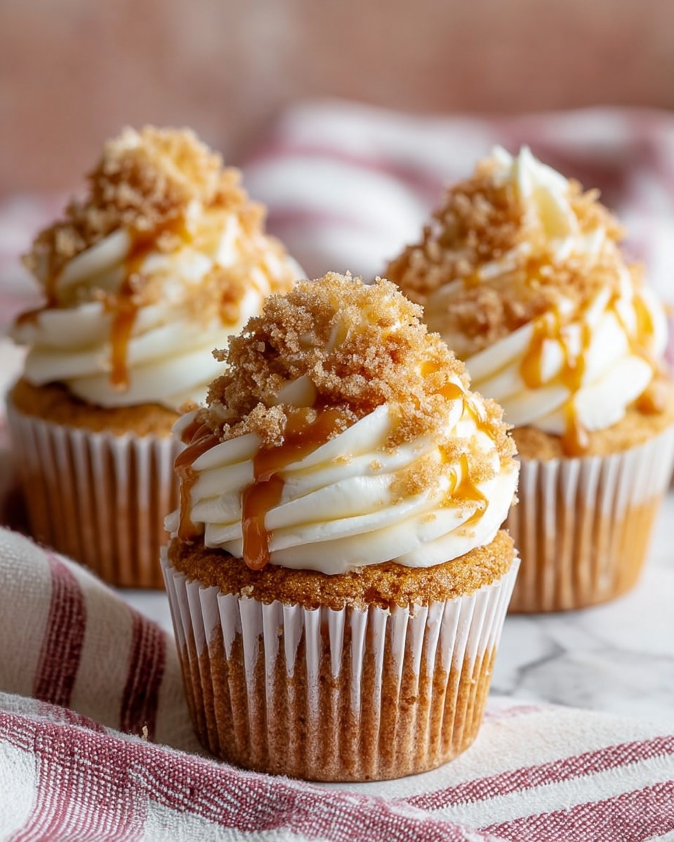 Three cupcakes are shown on a white marbled textured surface covered with a cloth featuring red and pink stripes. Each cupcake has one layer of golden brown cake wrapped in a white paper liner. On top of the cake is a thick swirl of creamy white frosting, shaped like soft waves. The frosting is covered with light brown crumbs sprinkled on top, and golden caramel sauce is drizzled over the crumbs and frosting, adding a shiny, sticky texture. The background is softly blurred with a warm pinkish tone. Photo taken with an iphone --ar 4:5 --v 7