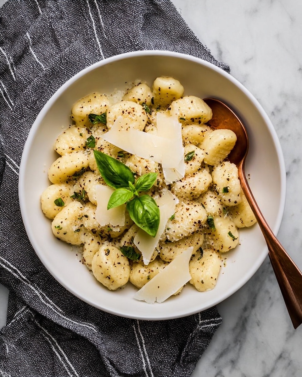 A white bowl filled with small, soft gnocchi pieces coated in a light, creamy sauce with specks of black pepper and green herbs scattered throughout. On top, thin, wide shavings of pale yellow cheese are layered in the center, garnished with two fresh green basil leaves. A copper-colored spoon rests inside the bowl on the right side. The bowl sits on a white marbled surface with a gray cloth featuring white stripes partially underneath it. photo taken with an iphone --ar 4:5 --v 7