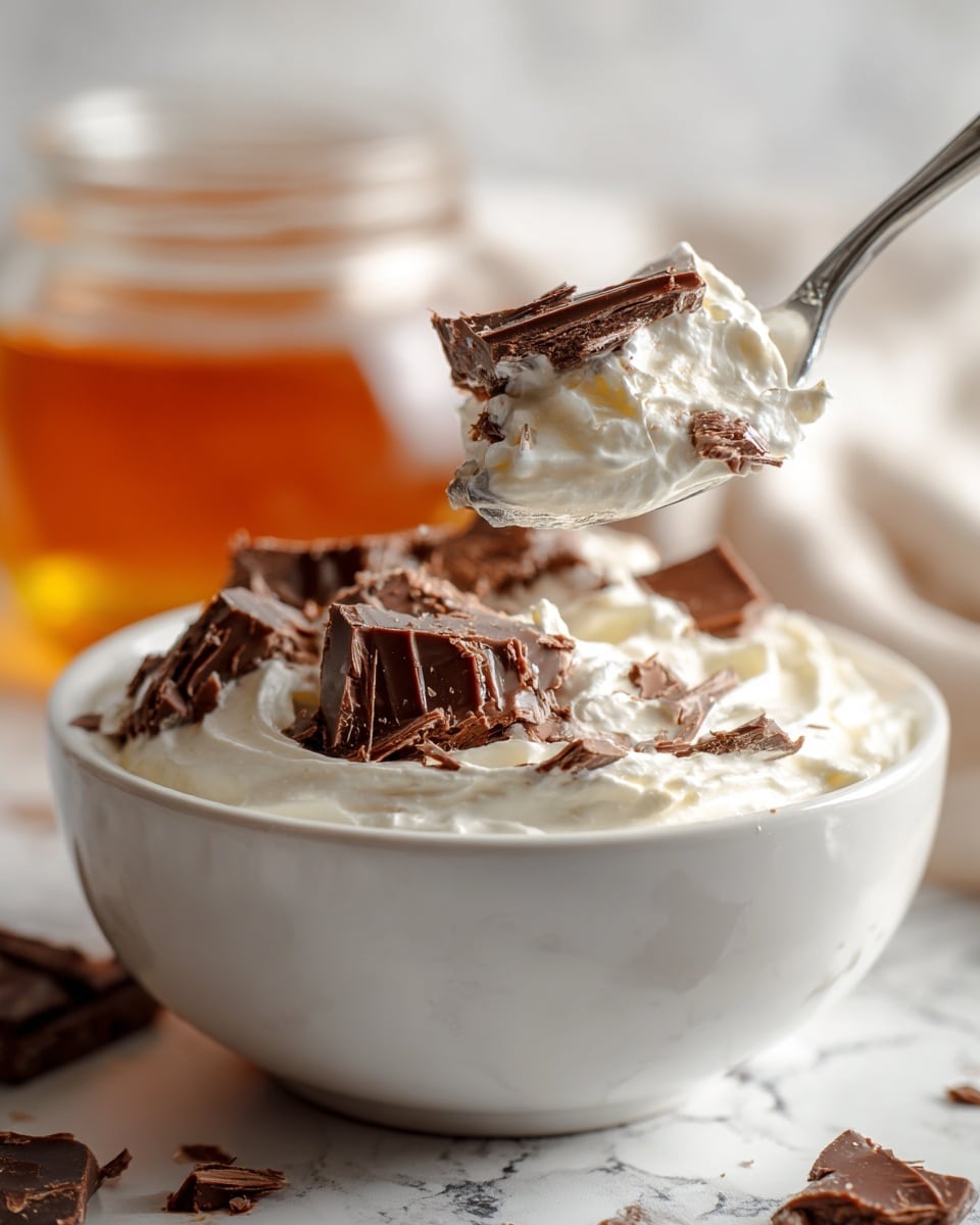 A close-up of a white bowl filled with a creamy, thick white layer mixed with broken pieces of solid milk chocolate that show a smooth, slightly bumpy texture. A silver spoon holds a scoop with a large chunk of chocolate on top, surrounded by the creamy white layer that appears soft and whipped. The background is blurred with a white marbled texture, and a glass jar filled with amber liquid sits behind the bowl. The overall image has a fresh and rich feel, showing contrast between the smooth white cream and the solid chocolate pieces. Photo taken with an iphone --ar 4:5 --v 7