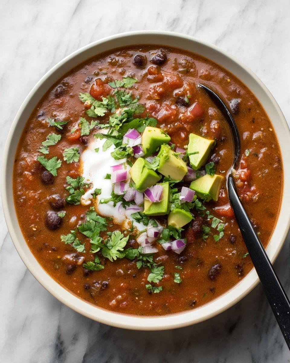 A white bowl filled with thick, reddish-brown bean soup that has visible black beans and small pieces of tomato throughout. On top, there is a dollop of white cream in the center, surrounded by small green avocado cubes and chopped purple onions. Fresh green cilantro leaves are scattered evenly across the soup. A black spoon rests inside the bowl, partially submerged in the soup. The bowl sits on a white marbled surface. photo taken with an iphone --ar 4:5 --v 7