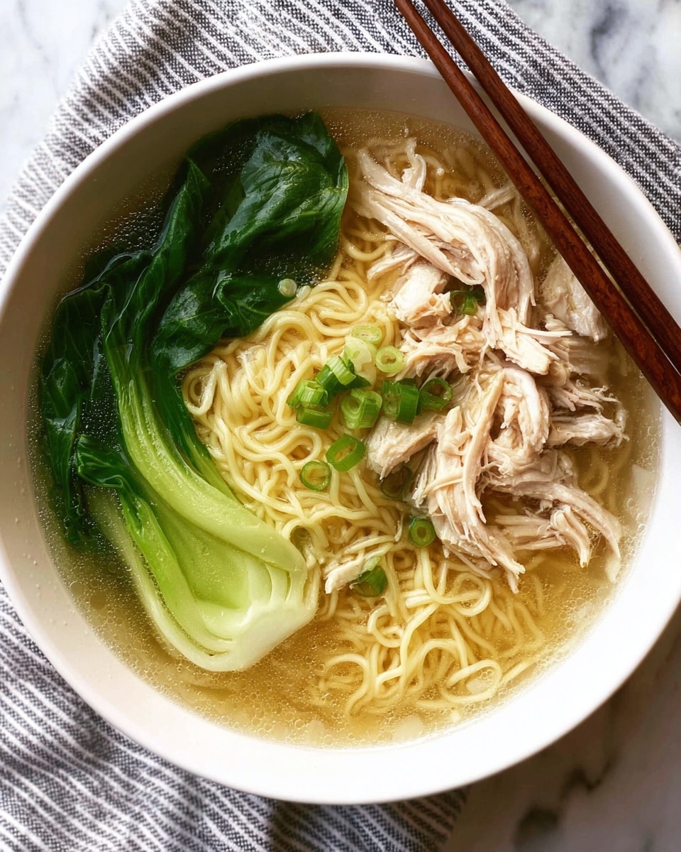 A white bowl filled with clear broth containing a nest of light yellow noodles at the center. On the left side, there are two bright green bok choy leaves with visible veins and a slightly shiny texture. On the right side, there are pieces of shredded white chicken meat layered loosely over the noodles. Small green onion slices are scattered over the noodles near the middle. A pair of brown chopsticks rests on the top right edge of the bowl. The bowl sits on a white marbled surface with a striped cloth underneath. Photo taken with an iphone --ar 4:5 --v 7