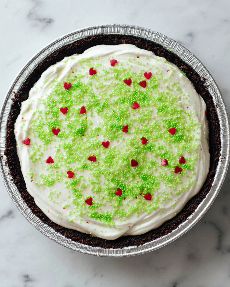 A round pie with a dark brown crumb crust forms the base and edges, held in a shiny silver pie tin. On top, a thick layer of smooth, white creamy filling spreads evenly with slight swirls. Bright green sugar sprinkles are scattered across the white layer, covering it almost like a light dusting. Small, glossy red heart-shaped sprinkles are also spread evenly over the top, giving a pop of color. The pie sits on a white marbled texture surface. photo taken with an iphone --ar 4:5 --v 7