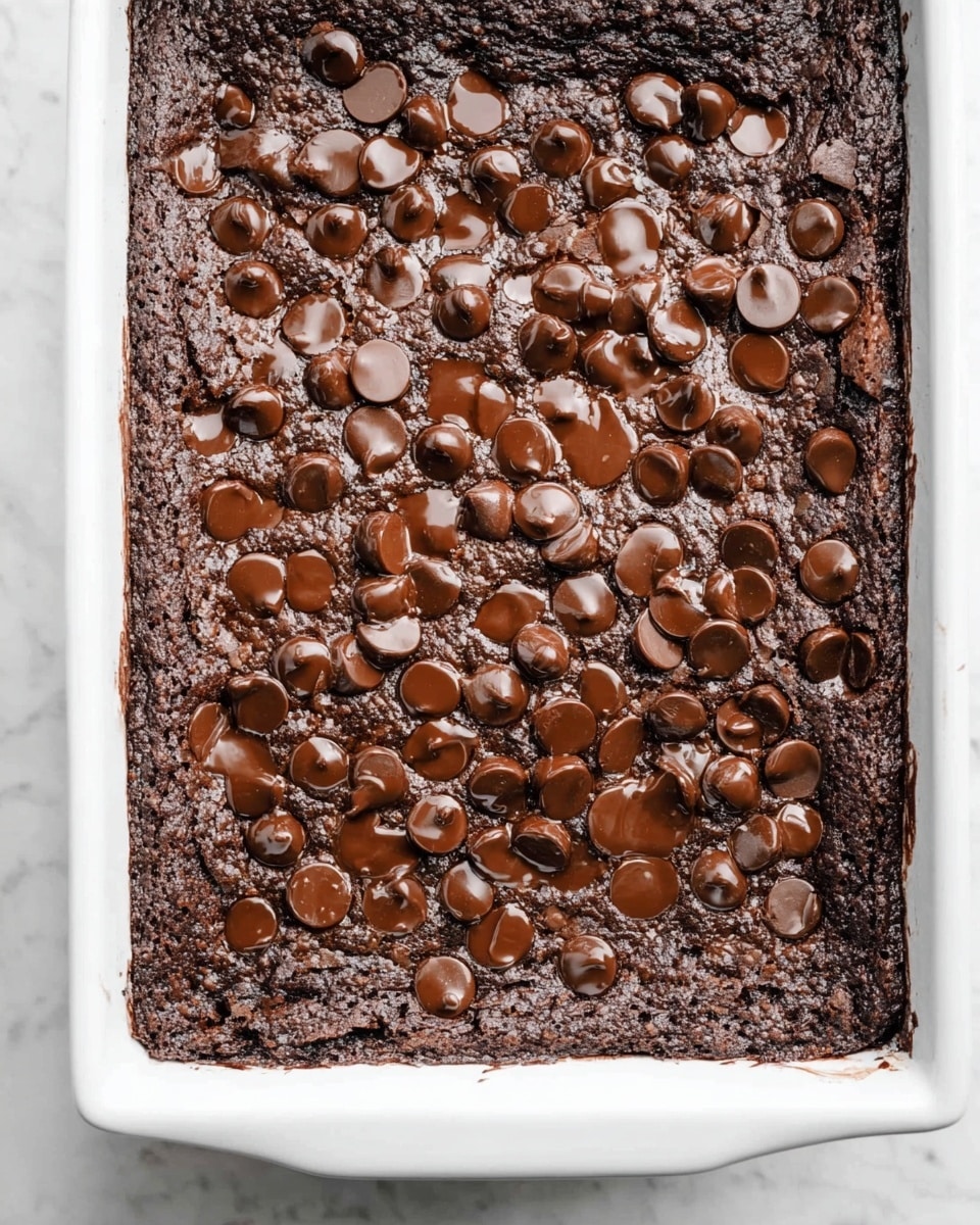 A baked chocolate brownie with a dark, cracked, and slightly rough texture forms the base layer, filling a white rectangular baking dish. The top layer is covered evenly with numerous glossy, melted chocolate chips that have softened and created small pools of shiny chocolate all over. The edges are slightly raised and appear crispier and denser compared to the moist center. The background is a white marbled texture. photo taken with an iphone --ar 4:5 --v 7