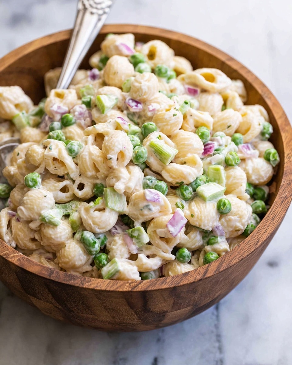 A wooden bowl filled with creamy pasta salad, showing about three layers of small shell pasta coated in a white creamy dressing, mixed with bright green peas, small chopped pale green celery pieces, and bits of red onion scattered evenly throughout. A silver spoon sticks out from the salad on the left side. The bowl sits on a white marbled surface, creating a fresh and clean background with soft natural lighting. photo taken with an iphone --ar 4:5 --v 7