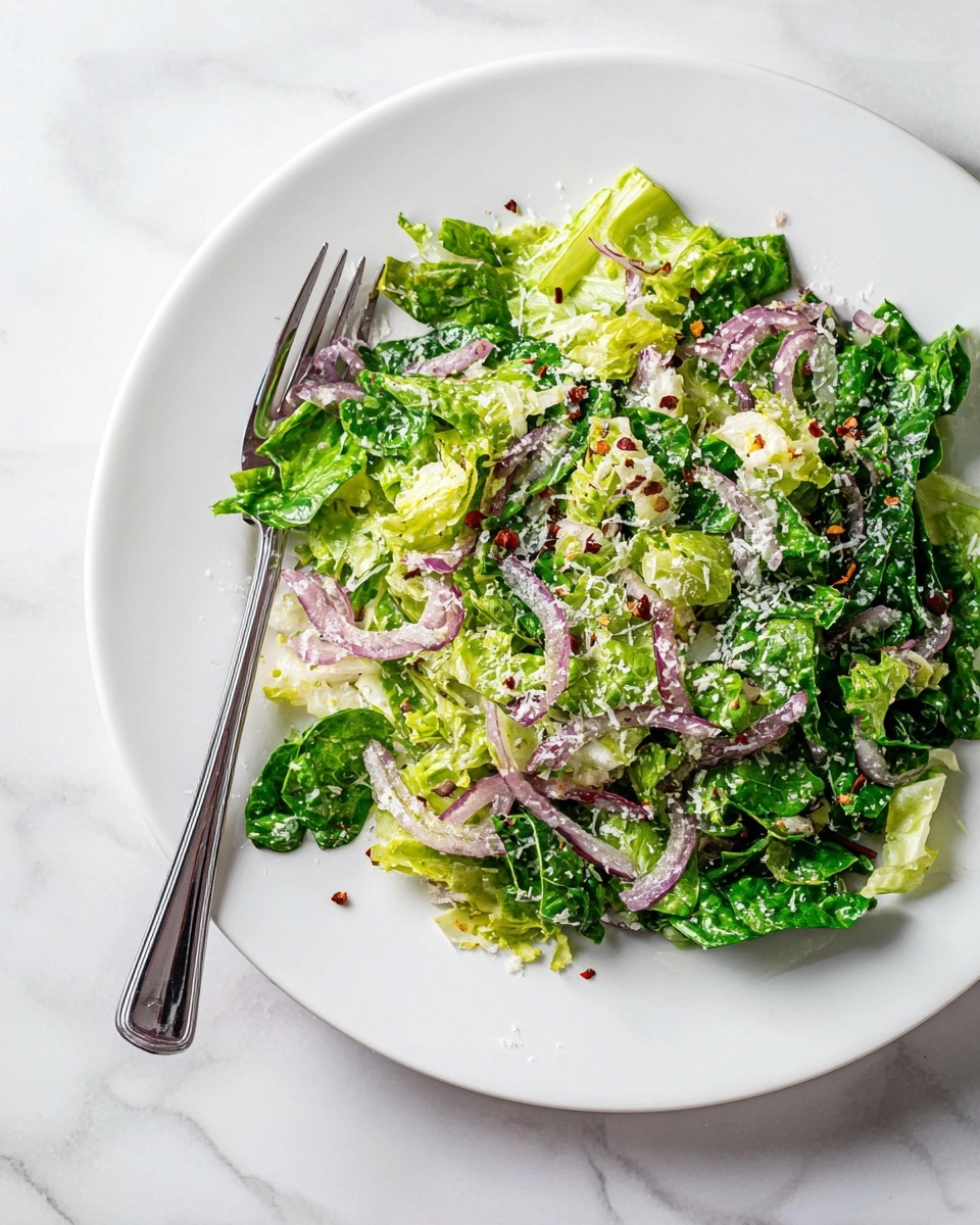 A white plate holds a fresh salad with two main layers: the base layer is made of bright green chopped leafy lettuce mixed with darker spinach leaves, giving a mix of light and dark green colors with a crisp texture. On top, thin slices of purple onion add a sharp color contrast and delicate crunch, scattered evenly throughout. The salad is sprinkled with fine white grated cheese and small red chili flakes that add texture and spots of color. A silver fork rests on the left side of the plate, and the whole scene sits on a white marbled surface. photo taken with an iphone --ar 4:5 --v 7