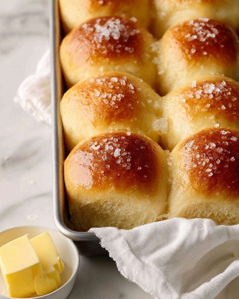 The image shows a close-up of nine soft, golden brown bread rolls baked closely together in a metal pan. Each roll is round with a shiny, slightly glossy top sprinkled with coarse salt crystals, creating a textured look. The rolls have a light, fluffy texture around the edges where they meet. Part of a white cloth around the pan is visible underneath, and a small white bowl with a stick of yellow butter sits nearby on a white marbled surface. The photo taken with an iphone --ar 4:5 --v 7