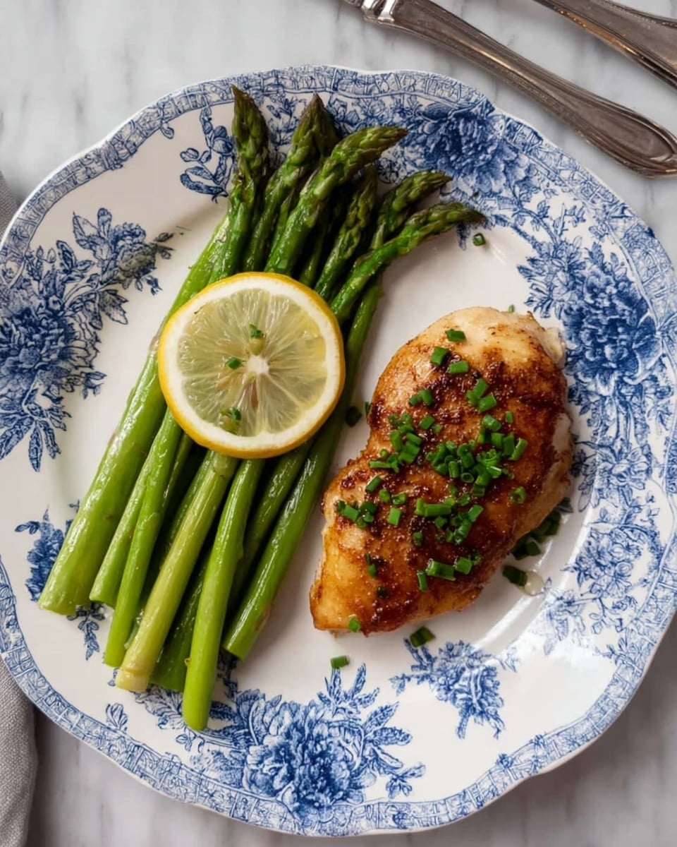 A white plate with blue floral patterns holds a meal consisting of two main parts arranged side by side; on the left, a bundle of bright green asparagus spears laid vertically topped with a lemon wedge angled downwards, and on the right, a golden-brown cooked chicken breast garnished with small chopped green herbs. The plate is placed on a white marbled surface, with a silver knife and fork visible near the top right edge. photo taken with an iphone --ar 4:5 --v 7