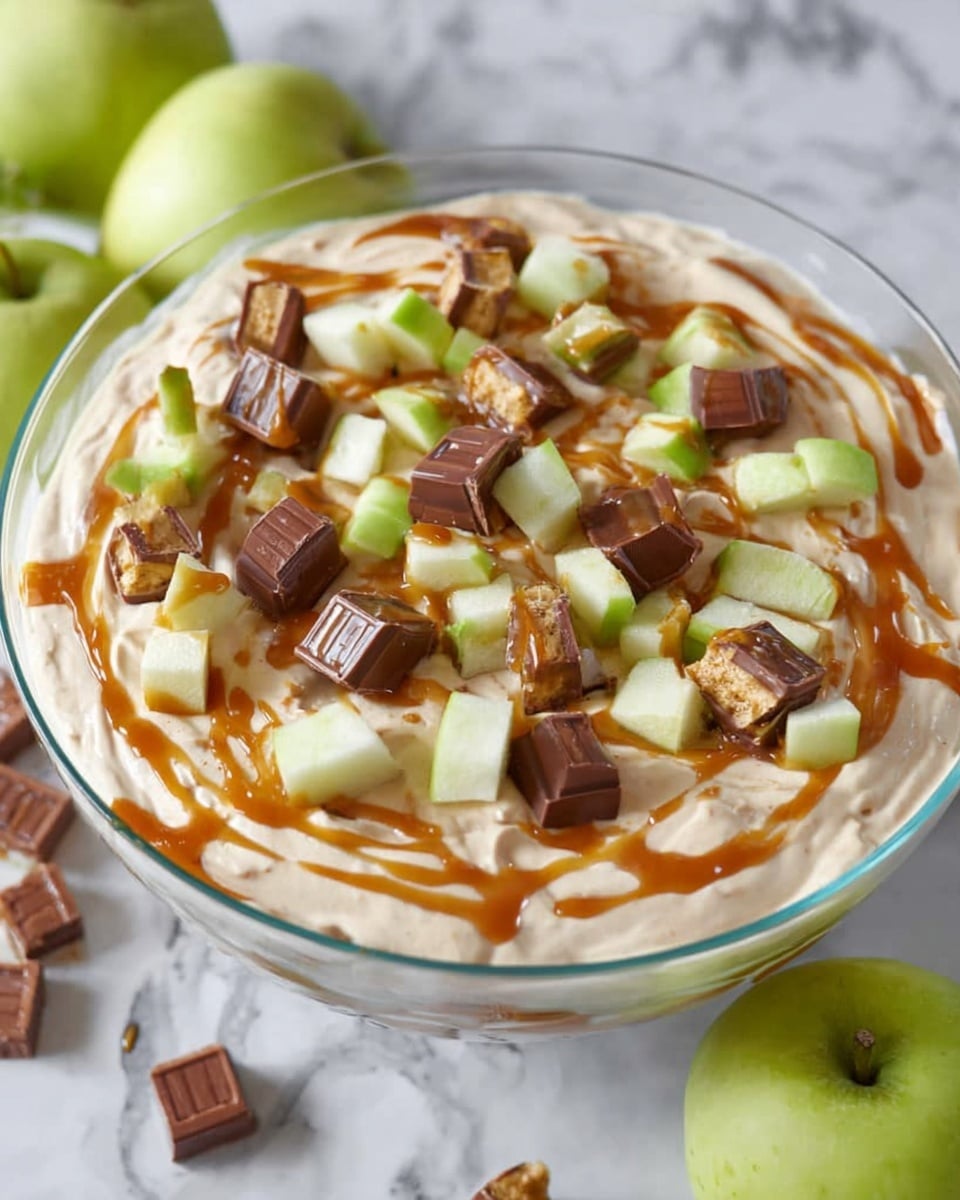 A clear glass bowl is filled with a creamy, light tan mixture with a smooth and fluffy texture, taking up the bottom and middle layers. On top, there are small chopped pieces of bright green apple with pale white insides, scattered evenly. Mixed among the apple chunks are small cubes of chocolate-covered candy bars showing layers of wafer and caramel inside. Drizzled over the top is a rich golden caramel sauce adding a glossy shine. The bowl sits on a white marbled surface with two whole green apples and some candy bars placed around it. Photo taken with an iphone --ar 4:5 --v 7