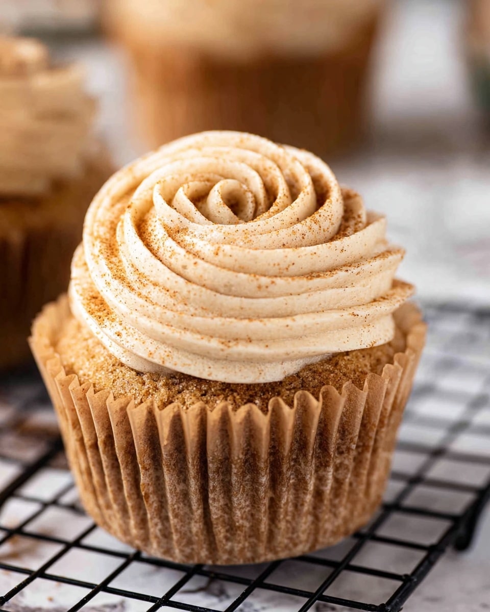 A close-up view of a single cupcake with a light brown, soft cake base, partially wrapped in a brown paper liner that is peeled back around the edges, showing its crumbly texture. On top, there is a thick swirl of creamy, light beige frosting with a smooth, slightly ridged texture, expertly piped in a circular motion. The frosting is lightly sprinkled with fine brown spice powder for decoration. The cupcake is placed on a black wire rack set against a white marbled surface, with another similar cupcake blurred in the background. photo taken with an iphone --ar 4:5 --v 7