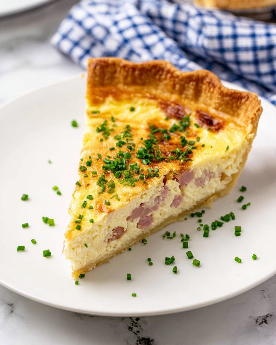A single slice of quiche sits on a white plate atop a white marbled surface. The quiche has three visible layers: a golden brown, flaky crust that forms a thick edge at the back, a creamy off-white middle layer with bits of pink ham embedded inside, and a slightly browned, smooth egg layer on top. Fresh chopped green chives are sprinkled over the top and scattered around the plate, adding a pop of color. The background includes a blue and white checkered cloth, blurred to keep focus on the quiche slice. Photo taken with an iphone --ar 4:5 --v 7