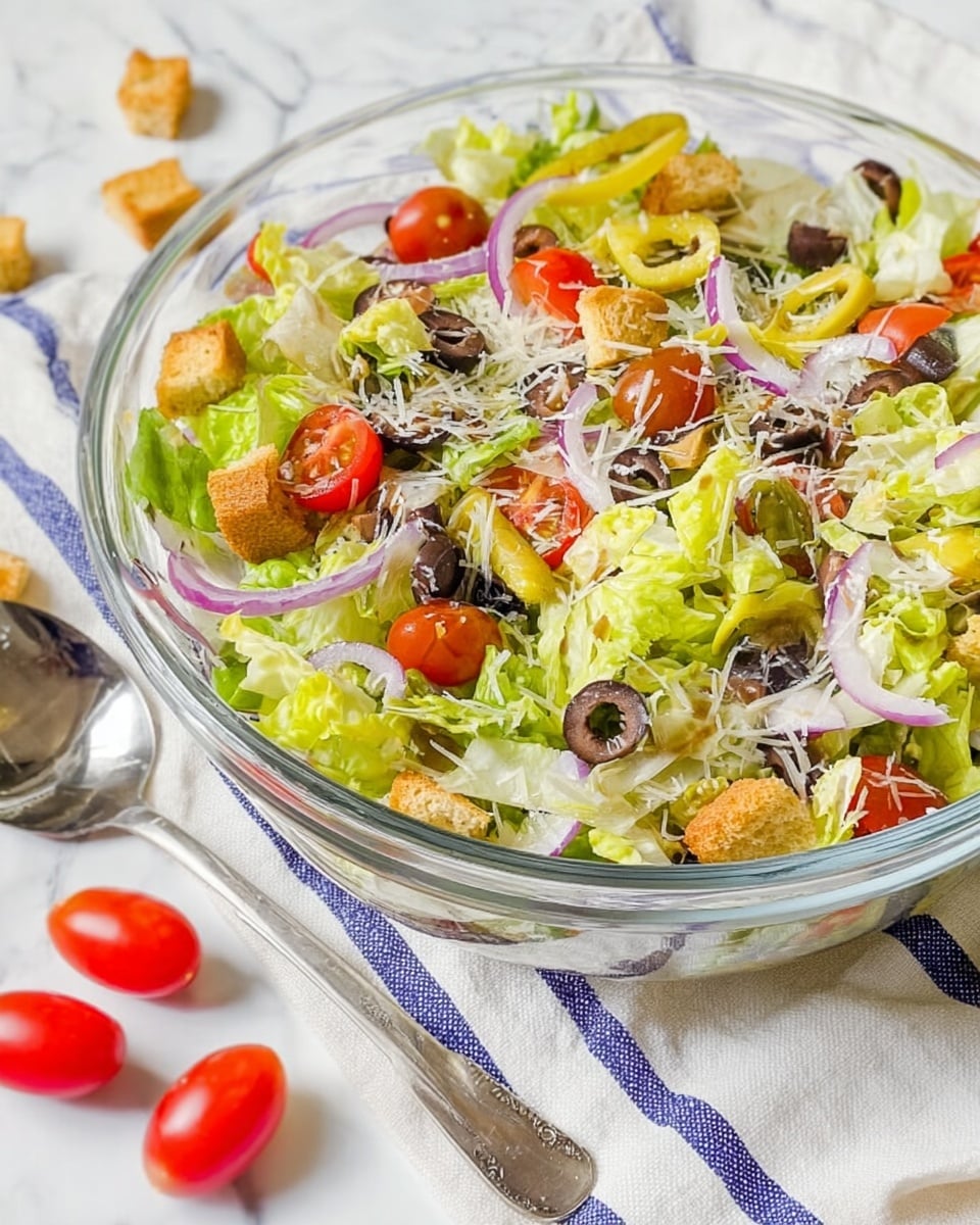 A clear glass bowl filled with a layered salad sits on a white marbled surface covered partly by a white and blue striped cloth. The salad has a base layer of bright green chopped lettuce, mixed with sliced red cherry tomatoes and thin rings of purple onions scattered throughout. Black olive slices and yellow pepper rings add contrast, while small golden brown croutons are spread on top. A fine sprinkling of shredded white cheese covers the surface, adding a light texture. A silver spoon rests near the bowl, with a few cherry tomatoes and croutons placed casually around it. Photo taken with an iphone --ar 4:5 --v 7