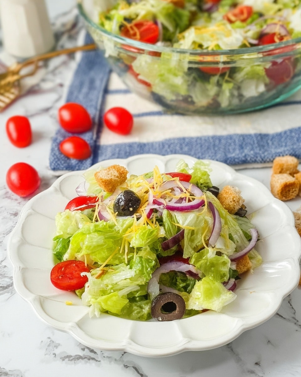 A fresh salad is served on a white scalloped plate with layers of light green lettuce, dark purple olive slices, thin red onion rings, bright red cherry tomato halves, and pale yellow shredded cheese on top, creating a colorful mix with varied textures. In the background, a clear glass bowl holds more of the same salad, placed on a white marbled surface with a blue and white striped cloth underneath. Red cherry tomatoes and small brown croutons are scattered around the plate, adding to the fresh and casual presentation, photo taken with an iphone --ar 4:5 --v 7