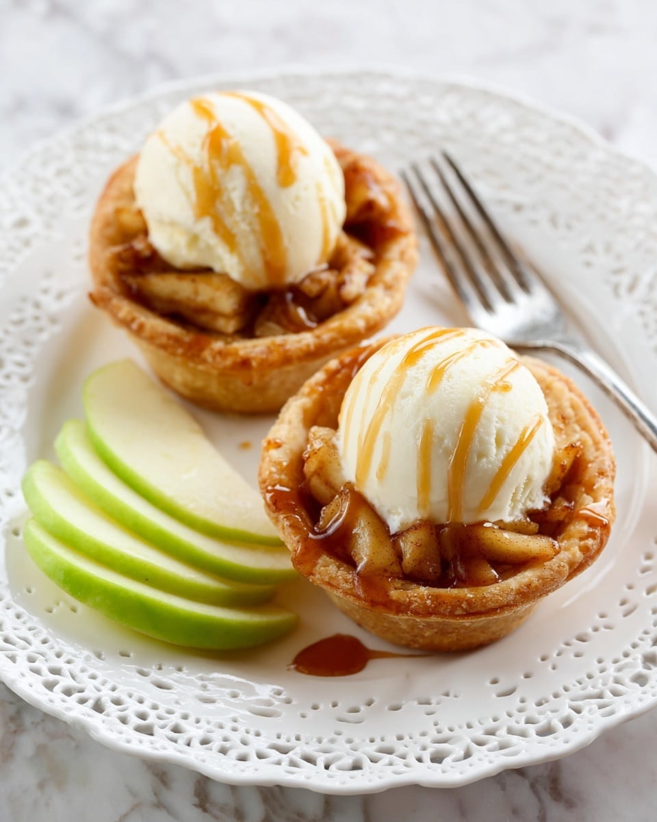Two small apple tarts rest on a white plate with a delicate lace-like edge, placed on a white marbled surface. Each tart has a golden-brown crispy crust forming a round cup holding a filling of warm, spiced apple pieces with a slightly caramelized texture. On top of each tart is a scoop of creamy vanilla ice cream with smooth, soft ridges, drizzled with a light amber caramel sauce that adds shine. To the side of the tarts, three thin slices of green apple fan out neatly. A silver fork lies on the plate near the upper tart, enhancing the inviting presentation. photo taken with an iphone --ar 4:5 --v 7