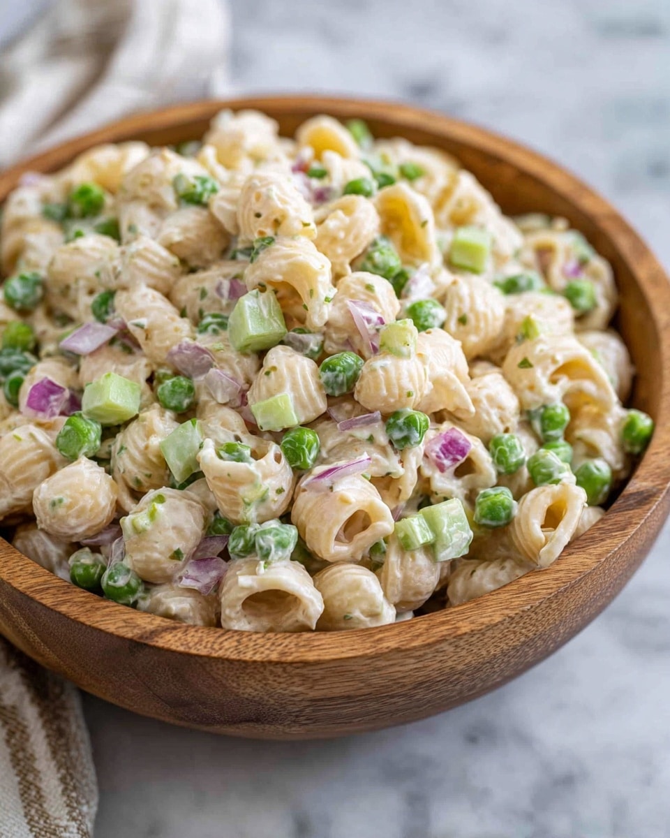 A wooden bowl filled with a creamy pasta salad, showing three main layers: the light beige pasta shells with a smooth texture form the base, mixed evenly with bright green peas and small cubes of pale green celery, both adding a fresh, crunchy look, while bits of purple-red onion are scattered throughout, providing small pops of color. The creamy dressing lightly coats everything, giving a slightly glossy finish. The bowl sits on a white marbled surface with a soft blurred background. photo taken with an iphone --ar 4:5 --v 7