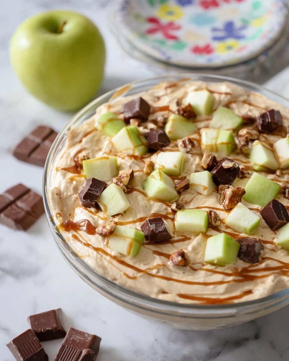 A clear glass bowl filled with a creamy beige mixture forms the base layer, topped with scattered pieces of light green apple cubes and small dark chocolate and nut candy chunks. A caramel sauce is drizzled lightly over the top, adding sticky golden lines across the surface. The bowl sits on a white marbled surface, with a whole light green apple and some chocolate candy pieces placed nearby. In the background, a white plate with a colorful floral pattern is partially visible. photo taken with an iphone --ar 4:5 --v 7