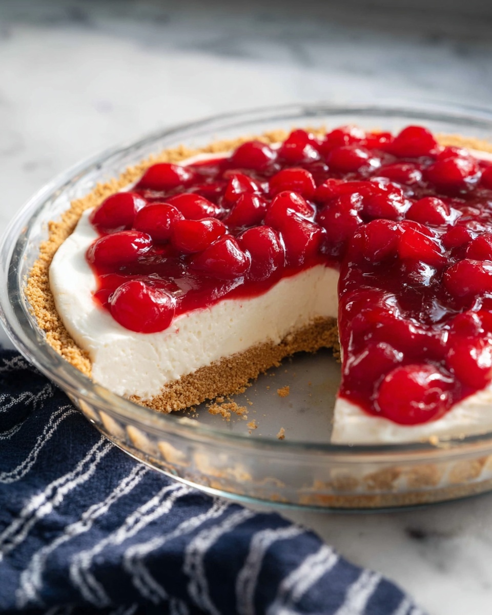 The image shows a round cherry cheesecake in a clear glass pie dish on a white marbled surface. The cheesecake has three visible layers: a bottom layer of crumbly, light brown graham cracker crust, a thick middle layer of smooth, creamy white cheesecake filling, and a top layer of bright red cherry topping with whole cherries covered in shiny glaze. A slice is cut out, revealing the clean edges of each layer. A navy blue cloth with white stripes is placed next to the pie dish. Photo taken with an iphone --ar 4:5 --v 7