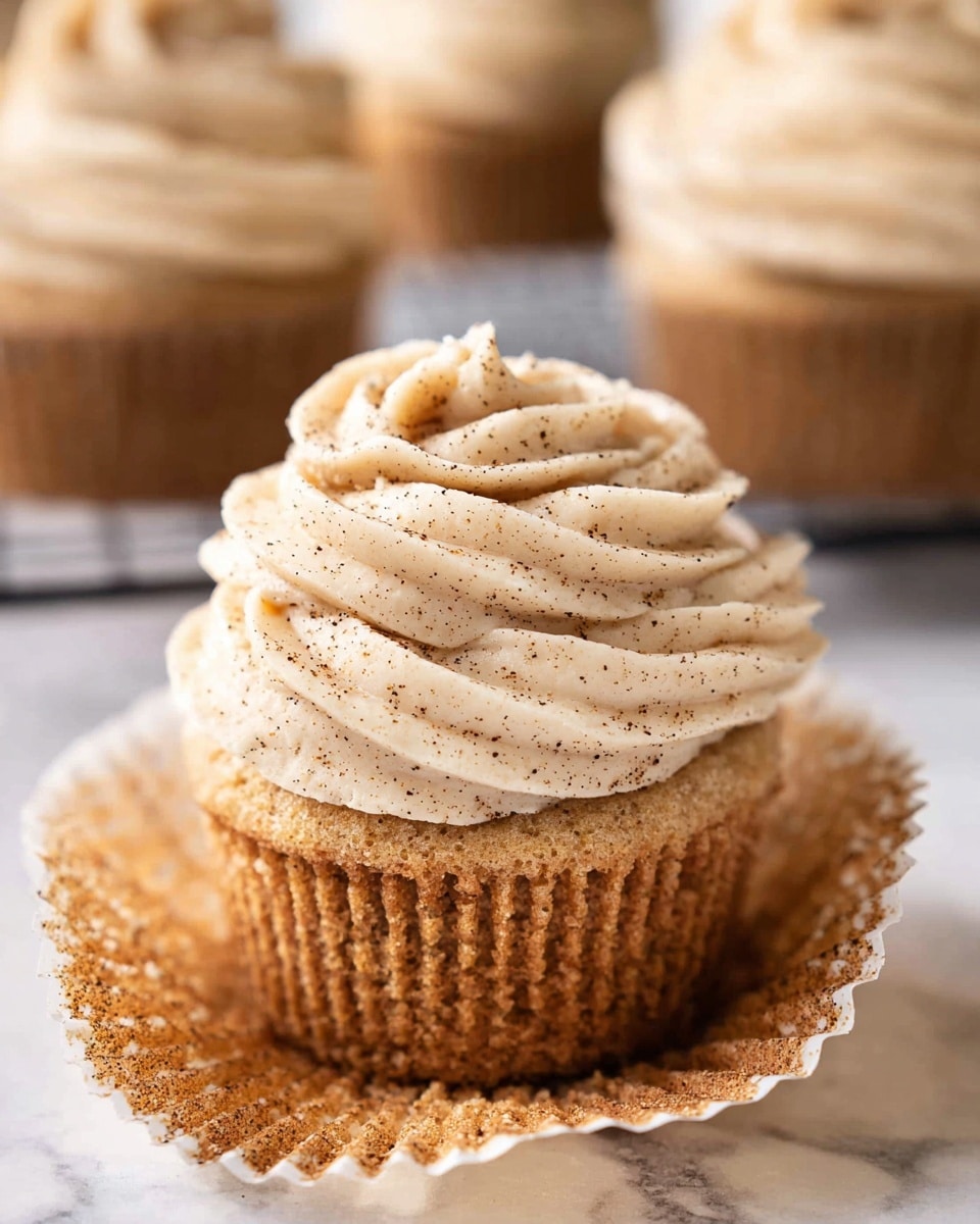 A close-up image of a single cupcake placed on a white marbled surface, partially unwrapped from its crinkled brown paper liner which reveals a light brown, soft base with a slightly crumbly texture. On top, there is a thick swirl of creamy frosting colored pale beige with a smooth and fluffy texture, sprinkled lightly with fine dark specks, possibly cinnamon or nutmeg, adding a touch of contrast. In the background, there are additional slightly blurred cupcakes with the same frosting style. The composition focuses on the detailed texture of both the cupcake base and the frosting. Photo taken with an iphone --ar 4:5 --v 7