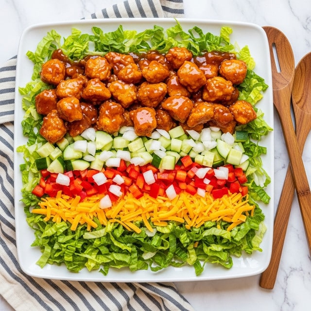 A white rectangular plate holds a layered salad starting with a base of bright green chopped lettuce spread evenly, followed by a colorful mix of chopped red bell peppers, pale cucumber cubes, and finely diced white onions scattered on top. The next layer is an orange shredded cheese sprinkled lightly in patches. The top layer consists of chunky, glazed, cooked chicken pieces coated with a shiny, dark orange sauce that covers most of the salad, creating a rich, textured look. The plate sits on a white marbled surface with a striped cloth and two wooden spoons placed nearby. photo taken with an iphone --ar 4:5 --v 7
