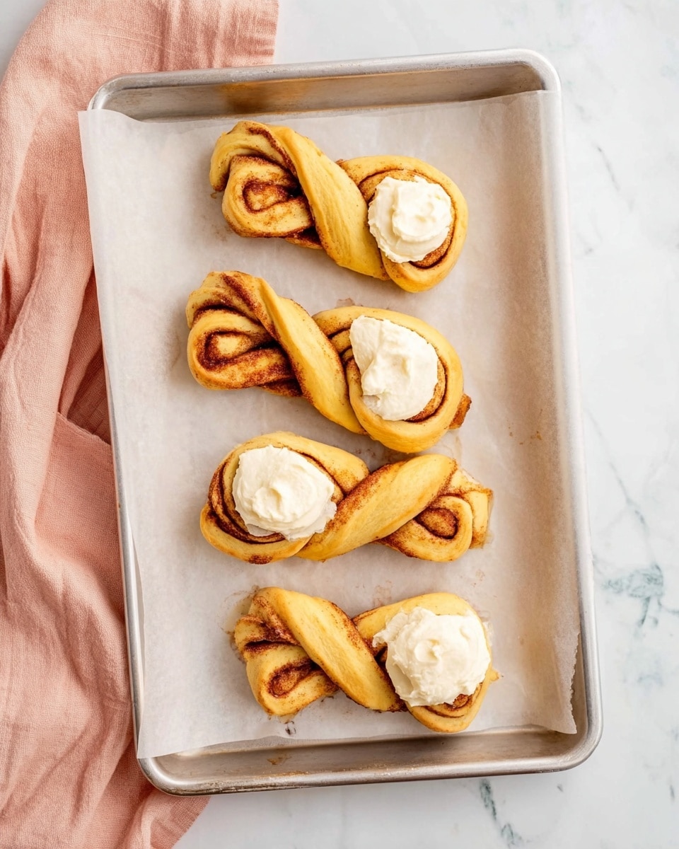 The image shows a silver baking tray lined with white parchment paper, holding five twisted cinnamon rolls before baking. Each roll has two visible layers of dough twisted together, showing a warm golden yellow color with darker brown cinnamon swirls. One end of each roll is topped with a dollop of creamy white frosting with a smooth, slightly swirled texture. The background under the tray is a white marbled surface, with a soft pink cloth partially visible beside the tray. Photo taken with an iphone --ar 4:5 --v 7