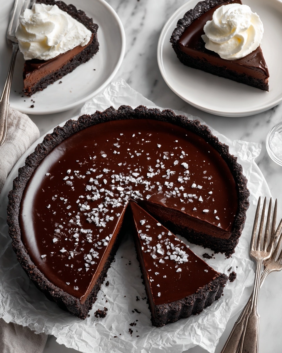 A round chocolate tart with three layers is shown on crumpled white parchment paper over a white marbled texture. The bottom layer is a dark, crumbly cookie crust, the middle is a lighter brown chocolate filling, and the top is a smooth, dark glossy chocolate ganache sprinkled with white flaky sea salt. One large slice is cut out from the tart, revealing all three layers clearly. Nearby, a white plate holds the removed slice topped with a dollop of white whipped cream, a silver fork is on the plate with a bit of tart on its prongs, and another fork is resting on a smaller white plate with a piece of tart. A clear glass bowl with coarse white sea salt is placed beside the tart. Photo taken with an iphone --ar 4:5 --v 7