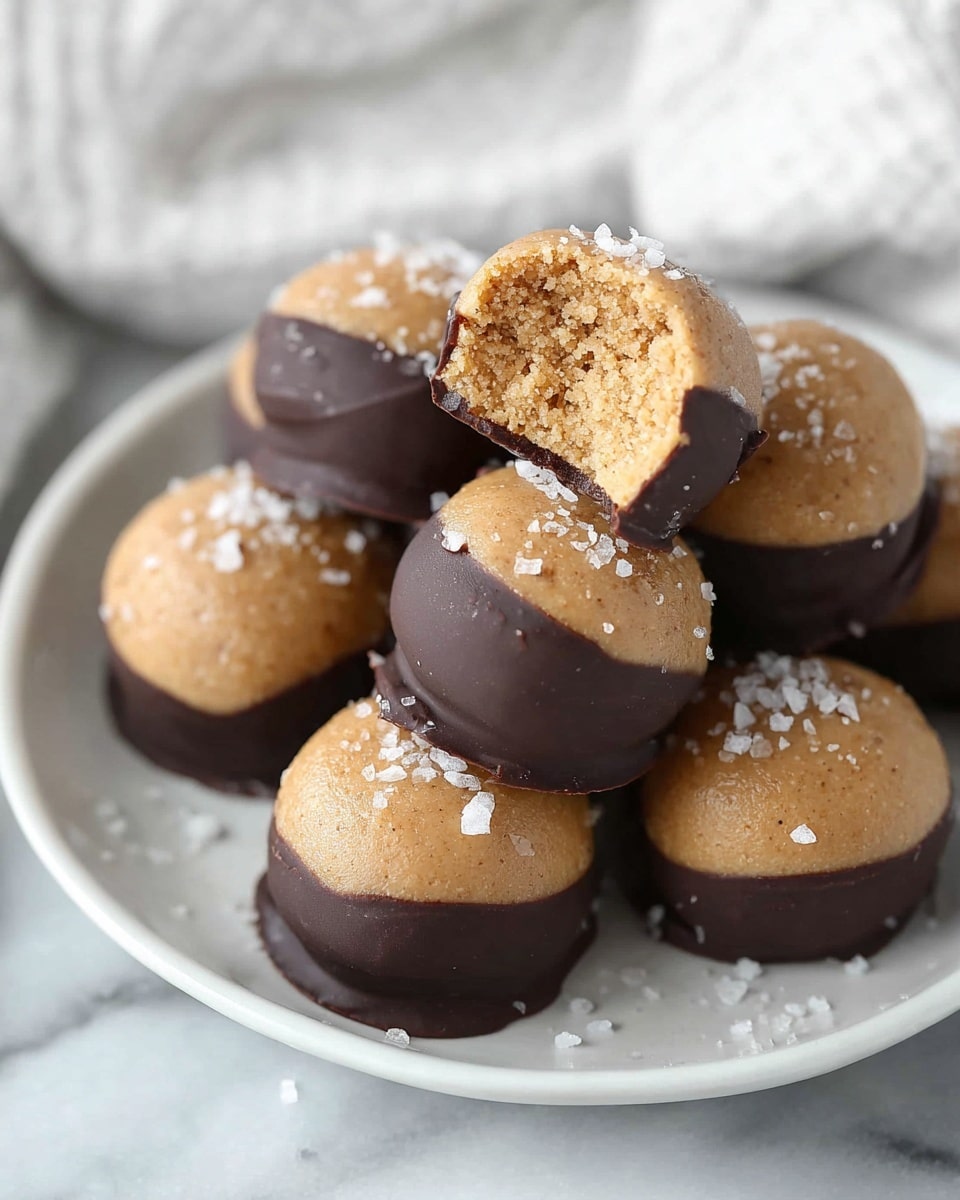 A white plate holds a pile of round treats, each with two clear layers: the top layer is light brown and smooth with a matte finish, while the bottom half is dipped in dark chocolate, creating a shiny, rich dark brown layer. The treats are sprinkled lightly with coarse white salt flakes on the light brown top. One treat is placed on top with a bite taken out, revealing a crumbly, dense, and slightly grainy light brown interior. The plate sits on a white marbled surface with a blurred white fabric in the background. photo taken with an iphone --ar 4:5 --v 7
