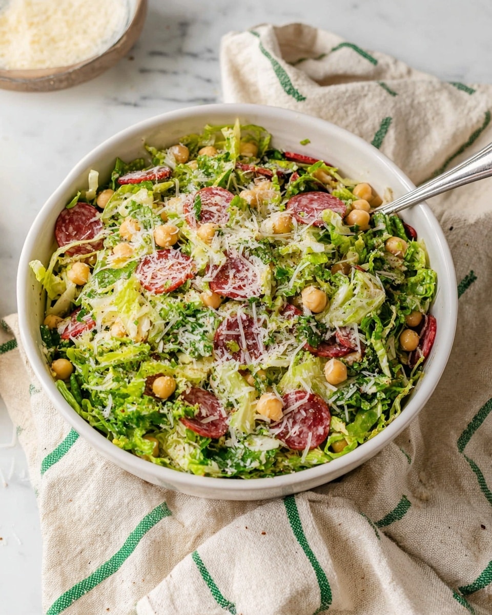 A white bowl filled with a fresh salad containing shredded green lettuce as the base layer, mixed with thin slices of dark red salami scattered throughout, and light beige chickpeas spread evenly. The salad is topped with a light dusting of grated white cheese, adding texture and creaminess. The bowl rests on a beige cloth with green and white stripes, all set on a white marbled surface. A metal spoon is placed inside the bowl on the right side. photo taken with an iphone --ar 4:5 --v 7