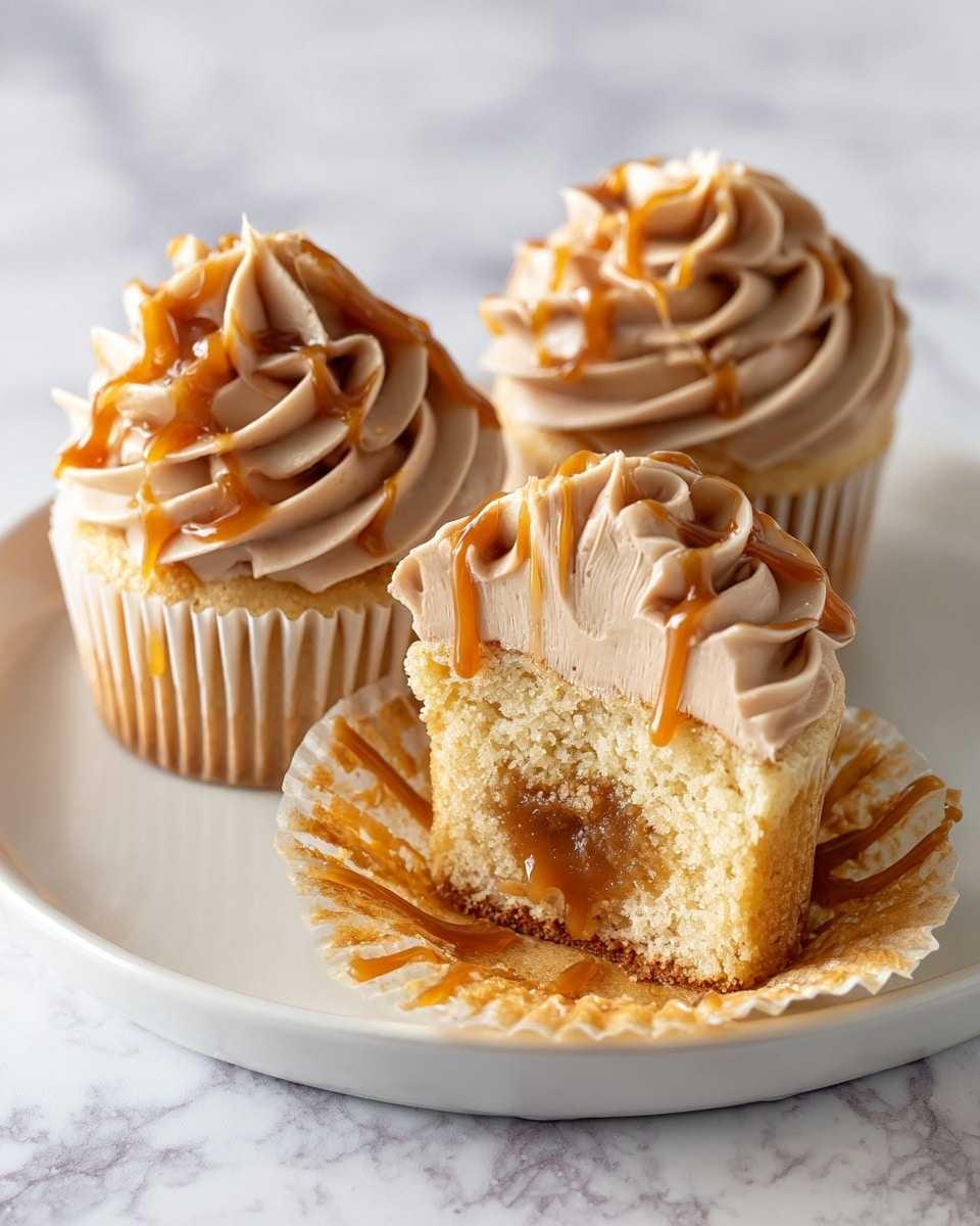 The image shows three cupcakes on a white plate, each topped with a thick swirl of light brown frosting that has a smooth and creamy texture. The frosting is decorated with drizzles of shiny caramel sauce that add a glossy accent. One cupcake is cut in half, revealing a soft yellow cake base with a gooey caramel center inside. The frosting on the cut cupcake is seen in layers on top, highlighting its fluffy texture. The setting is on a white marbled textured surface. Photo taken with an iphone --ar 4:5 --v 7