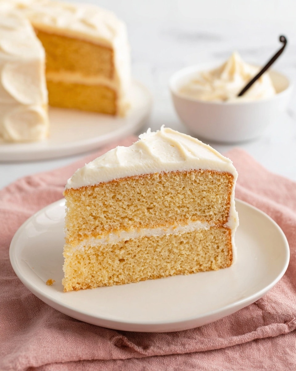 A slice of two-layer yellow cake sits on a white plate over a pink cloth on a white marbled surface. The cake layers are moist and spongy, with a light golden color and small air holes visible. Between the layers, there is a thin layer of smooth, creamy white frosting. The top layer is covered with a thick, even coat of the same white frosting, slightly glossy and softly textured with small swirls. In the background, a white bowl filled with extra frosting garnished with a vanilla bean and a white plate with more cake pieces are slightly blurred. photo taken with an iphone --ar 4:5 --v 7