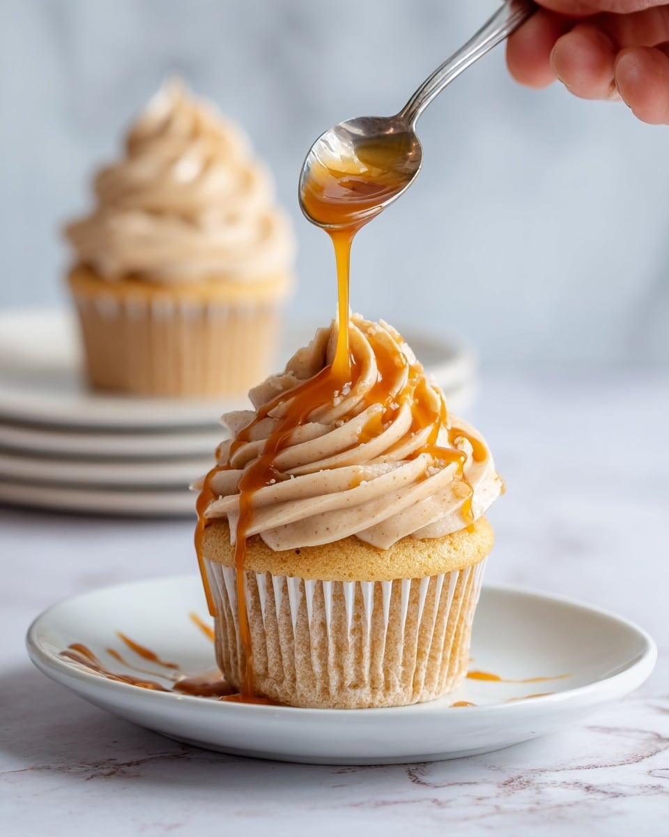 A yellow cupcake sits on a white plate with one thick swirl of light tan frosting on top, textured with soft, ruffled edges. A silver spoon held by a woman's hand drizzles golden caramel sauce over the frosting, with the caramel flowing down the layers and pooling slightly at the base. In the background, another cupcake with similar frosting can be seen on a white plate against a white marbled texture. The overall look is soft and inviting, with warm caramel tones contrasting the pale frosting and cake. Photo taken with an iphone --ar 4:5 --v 7