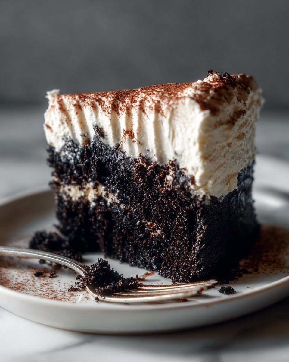 A close-up of a slice of dark, moist chocolate cake with a thick, creamy white frosting layer on top, slightly ridged with streaks of light brown powder dusted over the frosting. The cake sits on a white plate with a silver fork placed near the base, crumbs scattered around. The background is a smooth white marbled texture with soft lighting highlighting the cake's rich texture and creamy topping. photo taken with an iphone --ar 4:5 --v 7