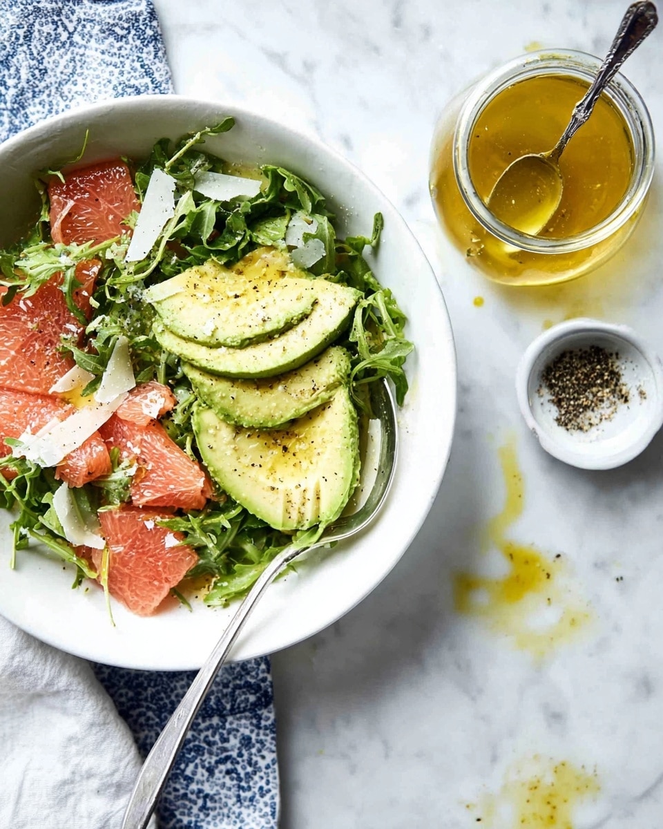 A white bowl filled with a fresh salad featuring three main layers: the base layer of green watercress leaves, medium-sized light green slices of avocado arranged on top, and bright pink grapefruit chunks scattered around. The salad is garnished lightly with thin white shavings of cheese. Next to the bowl, there is a small glass jar of golden olive oil with a spoon inside. Also visible is a small white container with black pepper and a blue patterned cloth on a white marbled surface. Some small splashes of oil and crumbs are spread around. Photo taken with an iphone --ar 4:5 --v 7