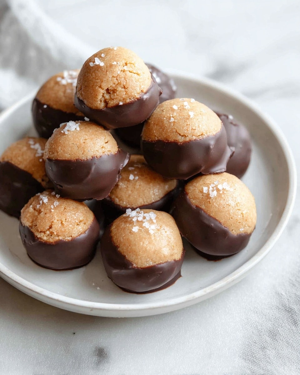 A small white plate holds a pile of round treats, each with two layers: the top half is light brown and slightly rough in texture, looking like a cookie or dough, while the bottom half is coated in smooth, dark chocolate that covers about half of each piece. Some of the treats are sprinkled lightly with coarse sea salt on top, adding a bit of texture and shine. The plate sits on a white marbled surface with soft, natural lighting highlighting the contrast between the chocolate and the cookie part. photo taken with an iphone --ar 4:5 --v 7