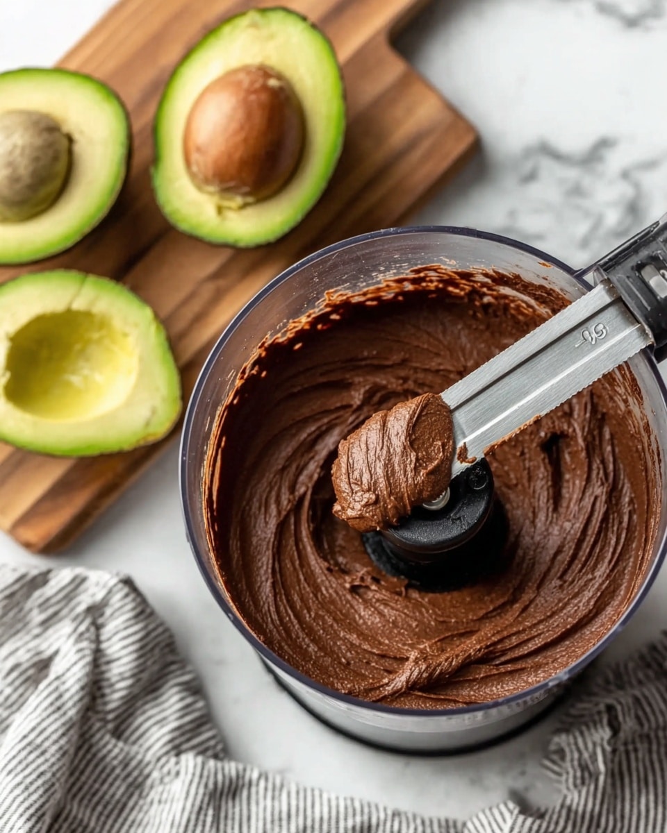 The image shows a close-up of a food processor filled with smooth, thick chocolate mousse that has a rich, dark brown color and creamy texture; the food processor blade is lifted slightly with some mousse clinging to it. To the left, there are two halved avocados with bright green flesh and large brown seeds, placed on a wooden cutting board. The whole scene is set on a white marbled surface with a grey and white striped cloth nearby. Photo taken with an iphone --ar 4:5 --v 7
