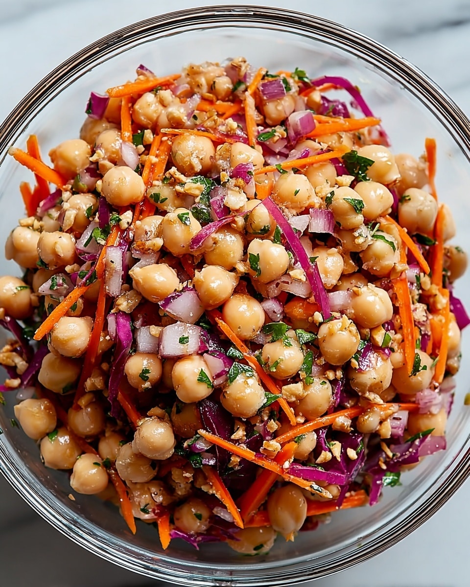 A clear glass bowl holds a chickpea salad with three main visible layers mixed together: beige round chickpeas as the base, thin bright orange carrot sticks scattered throughout, and chopped purple-red onions adding vibrant color. Small green parsley leaves are finely chopped and mixed evenly with the salad. The salad is lightly coated with a dressing that gives a slight shine to the ingredients, and some crushed nuts or seeds add a rough texture on top. The bowl sits on a white marbled surface, enhancing the fresh and colorful look of the dish. photo taken with an iphone --ar 4:5 --v 7