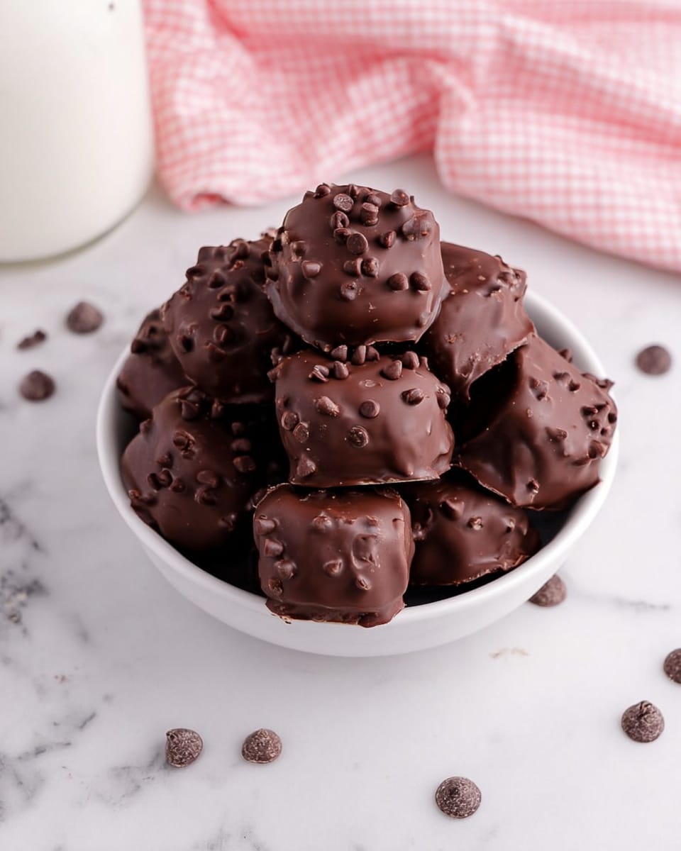 A white bowl is filled with round and square-shaped chocolate-covered treats, each covered in a thick layer of smooth dark chocolate with small chocolate chips sprinkled on top, giving a slightly rough texture. The treats are piled high, showing glossy, rich brown surfaces. Some tiny chocolate chips are scattered around the bowl on the white marbled surface. In the background, a white container and a pink checkered cloth add soft colors to the scene. Photo taken with an iphone --ar 4:5 --v 7
